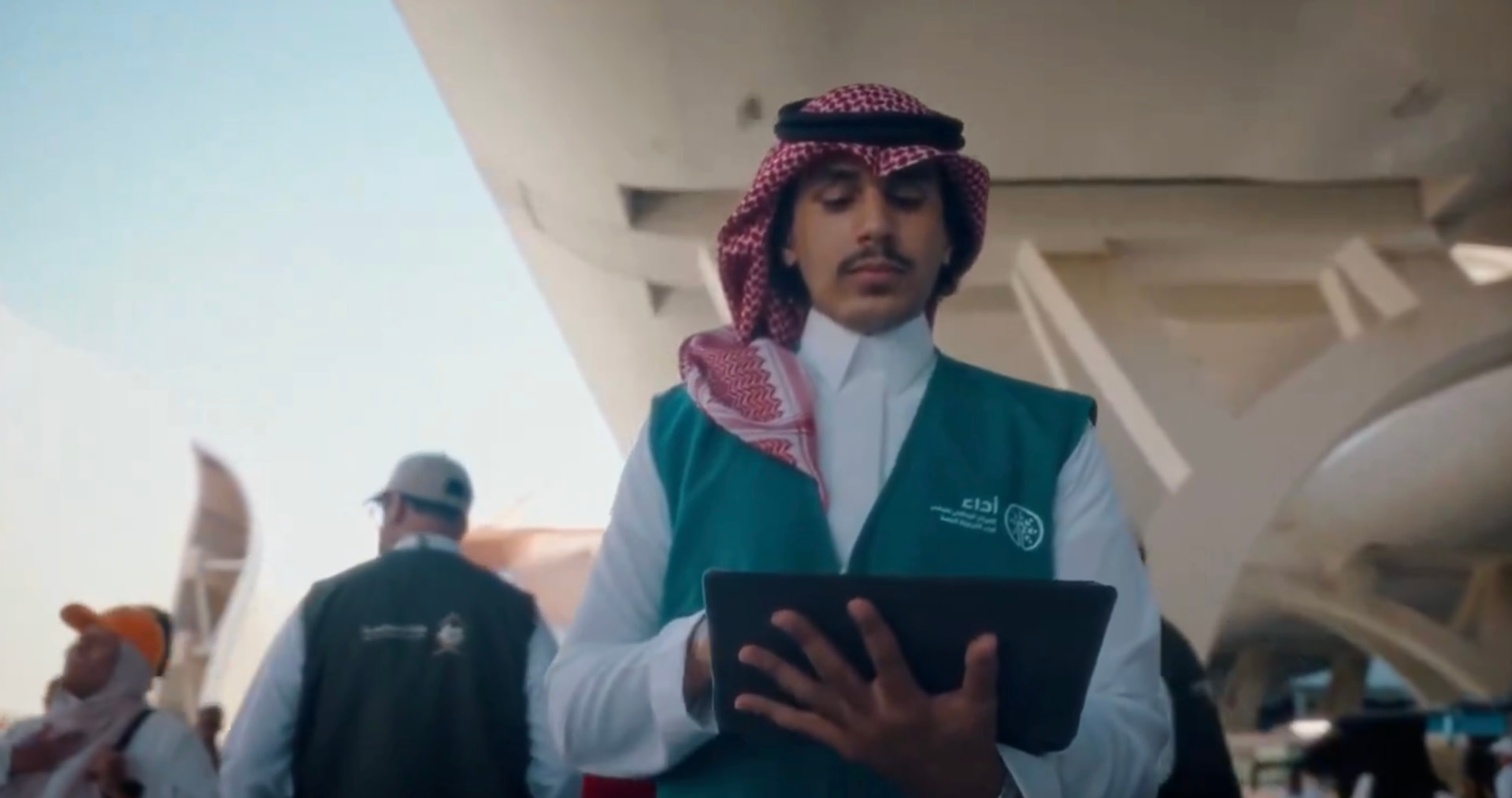 A young Saudi volunteer in a green Ada'a vest holding a tablet in the Hajj terminal — يسر و طمأنينة