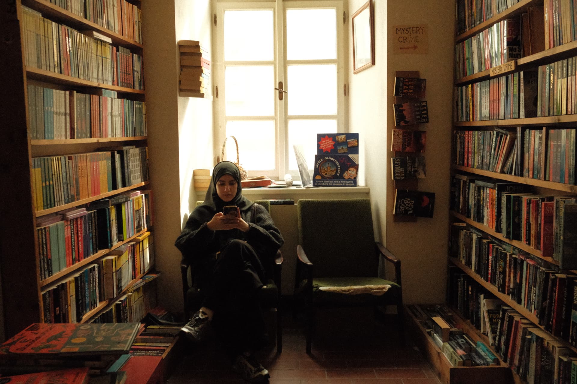 Fatima Mustafa seated in a green armchair in a bookshop, looking at her phone