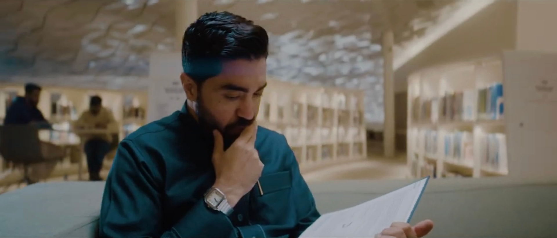 A man seated in a library reading an open folder — إثراء الإنسان